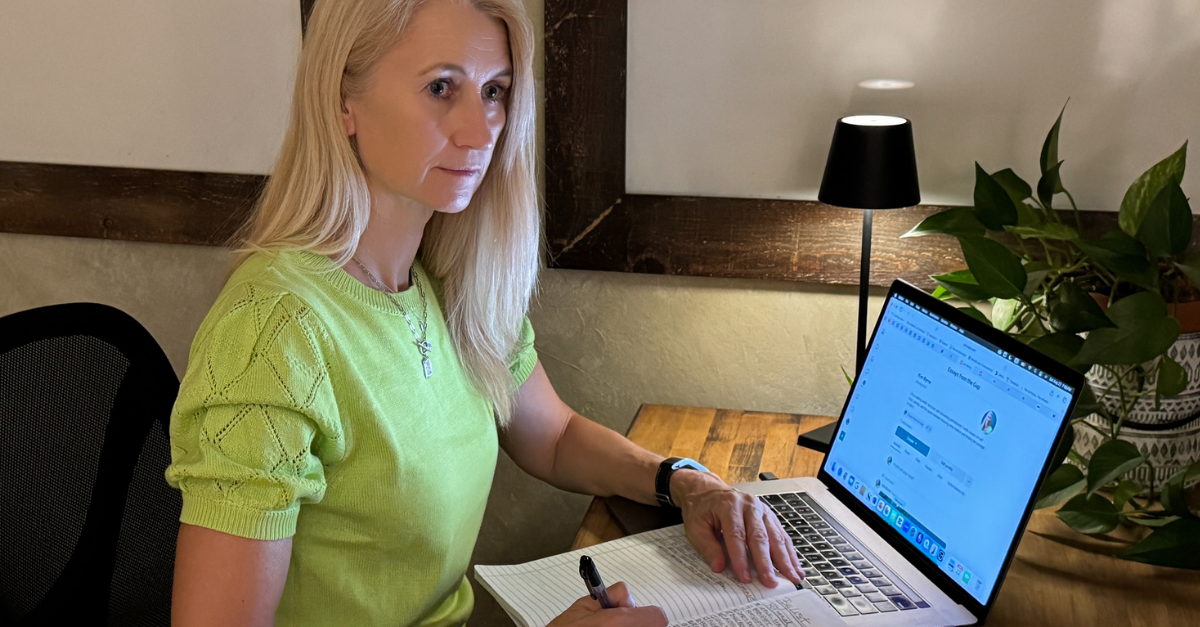 Kim Byrne sitting at her desk beside a laptop and handwritten checklist, looking away from the work in natural light.