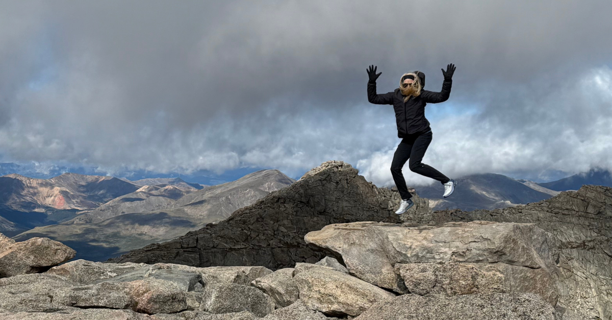 Kim Byrne jumping at the Mount Blue Sky summit marker in Colorado, bundled in layers on a cold, windy day.